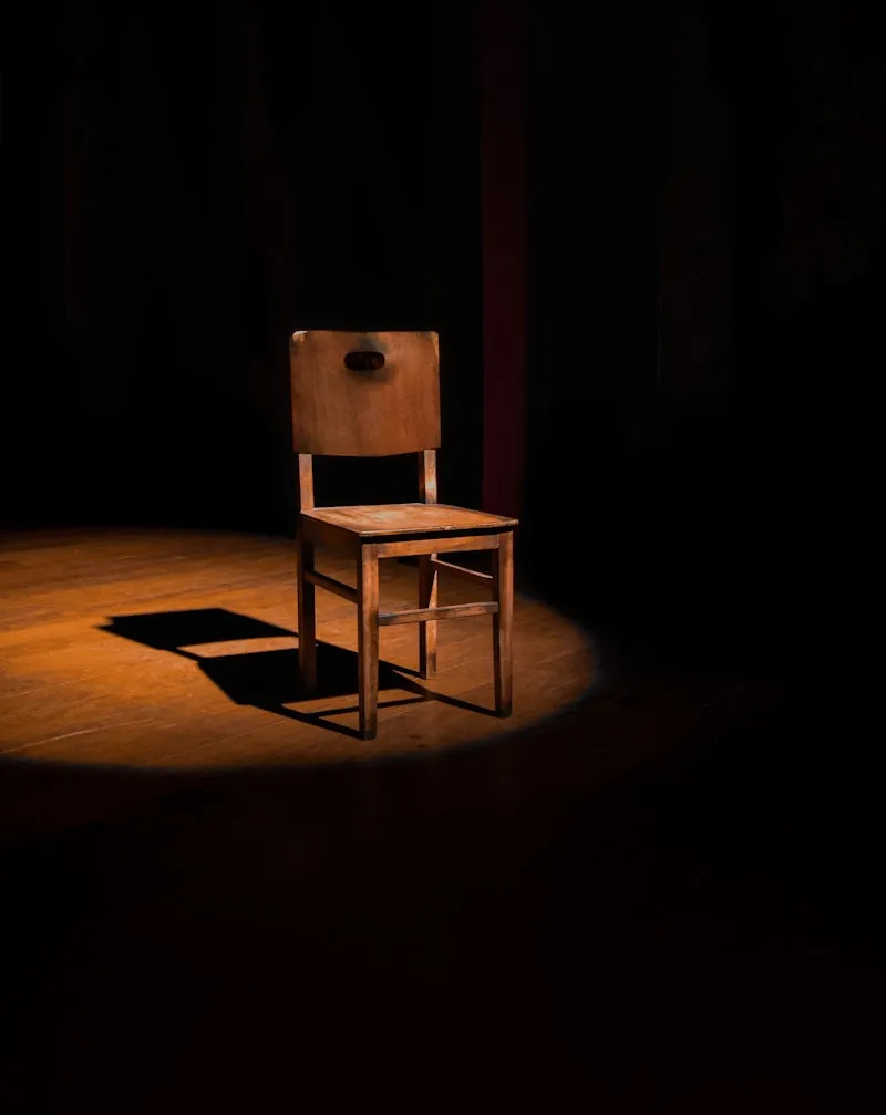 A simple wooden chair lit in a single spotlight on a dark stage.